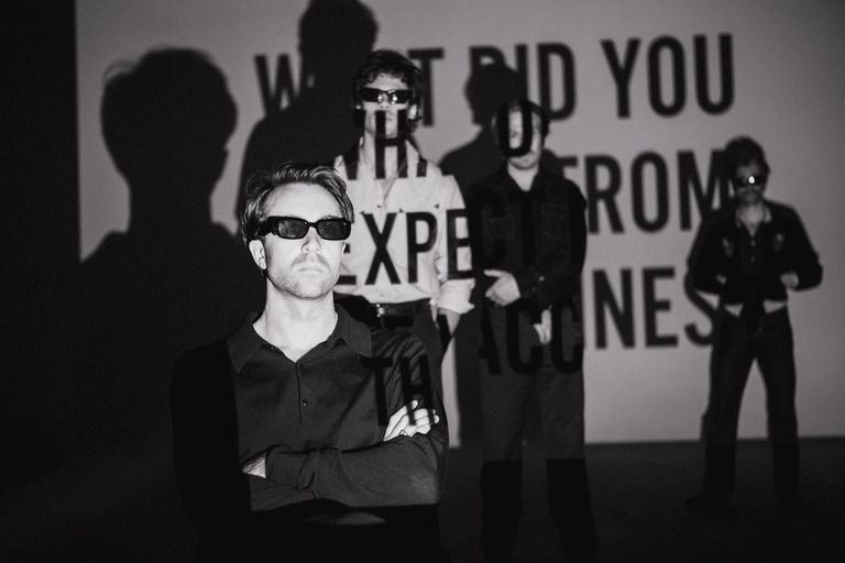 Indie rock band The Vaccines in a black-and-white portrait with projected text reading What Did You Expect From The Vaccines.